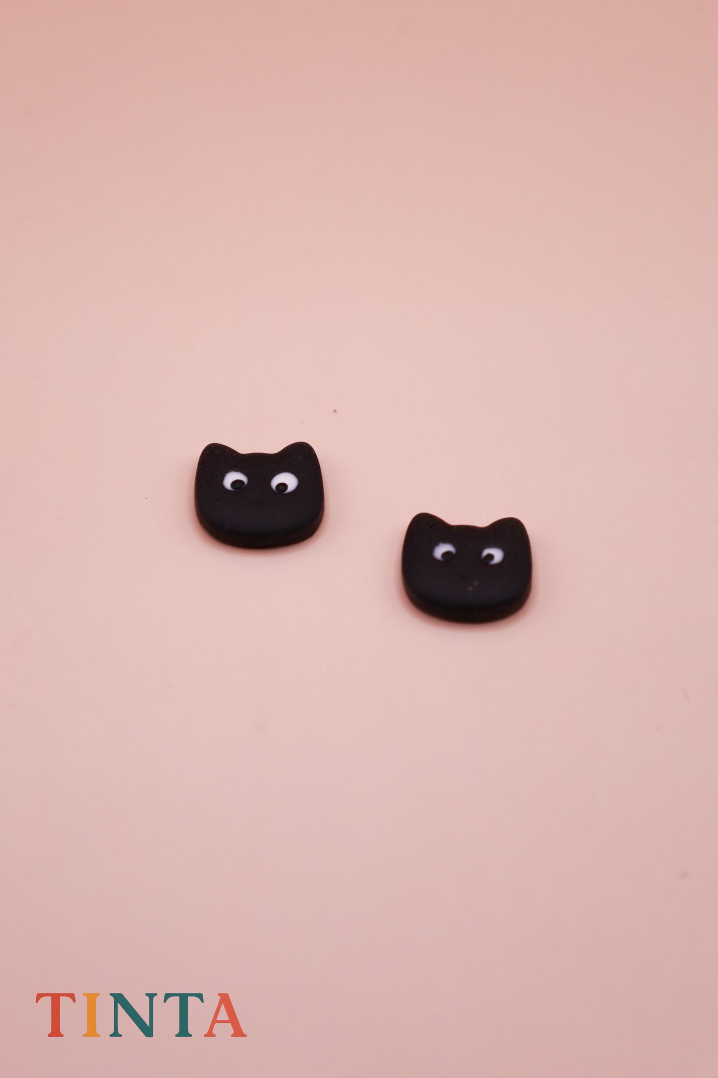 Black cat-shaped earrings on a pink background with 'TINTA' branding.
