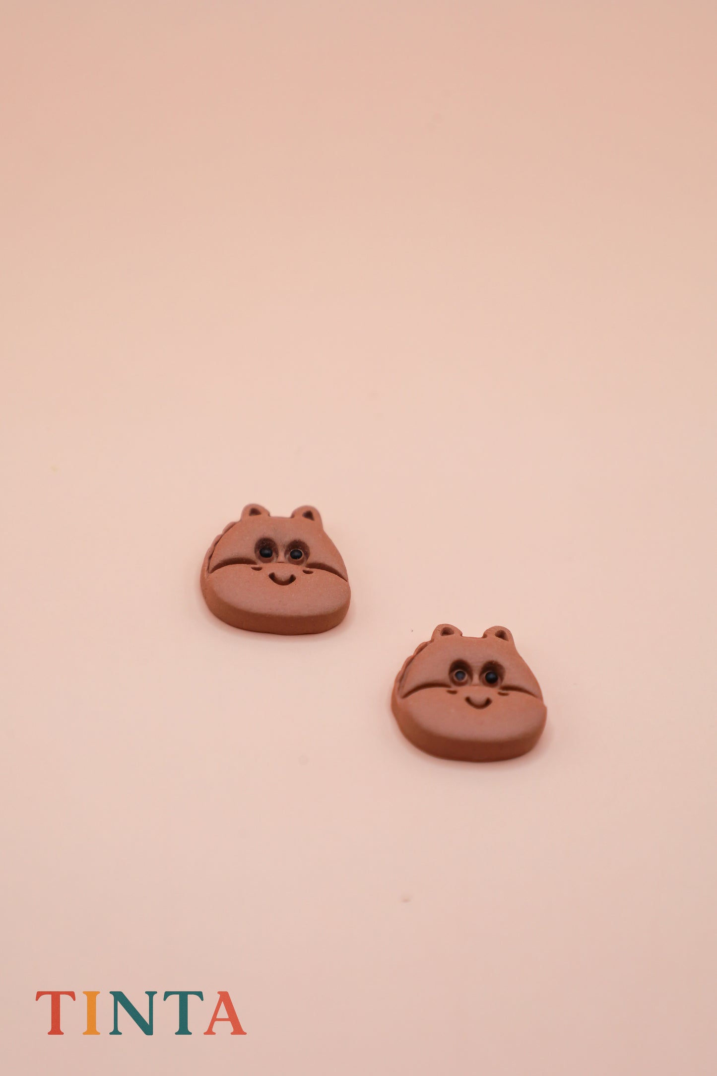 Two brown cat-shaped earrings on a beige background with 'TINTA' branding.