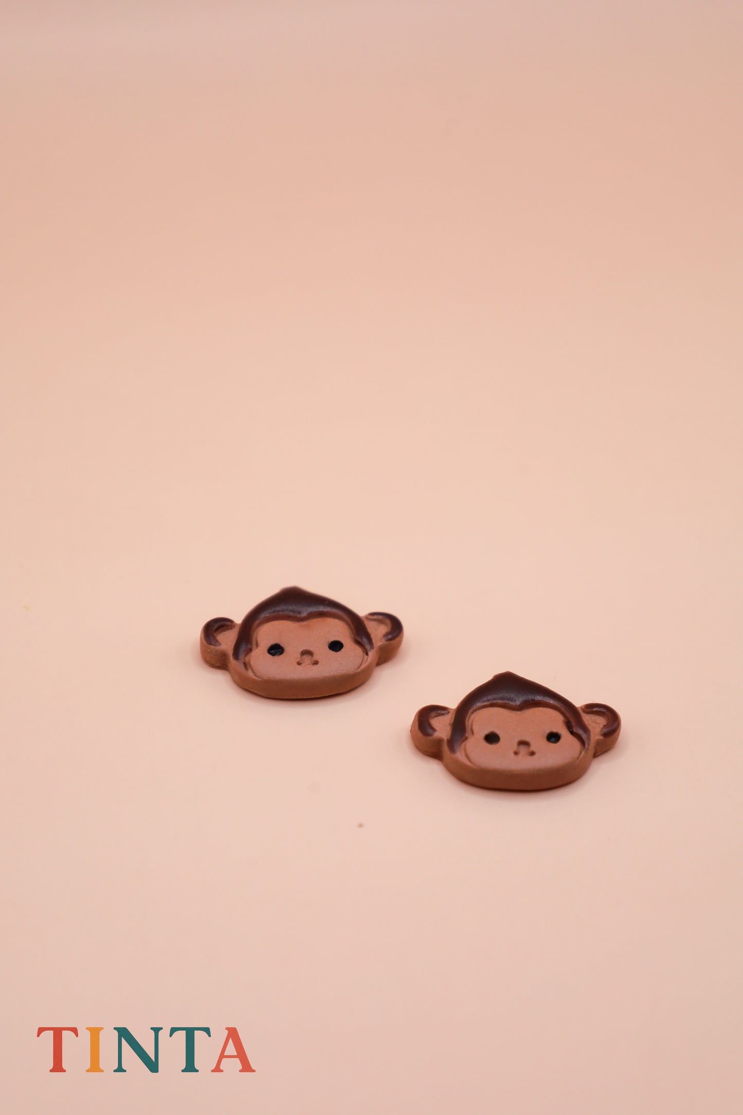 Two monkey-shaped earrings on a beige background with 'TINTA' branding.