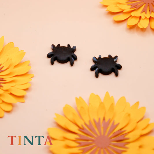 Black spider earrings with yellow flowers on a beige background, featuring the brand 'TINTA'.
