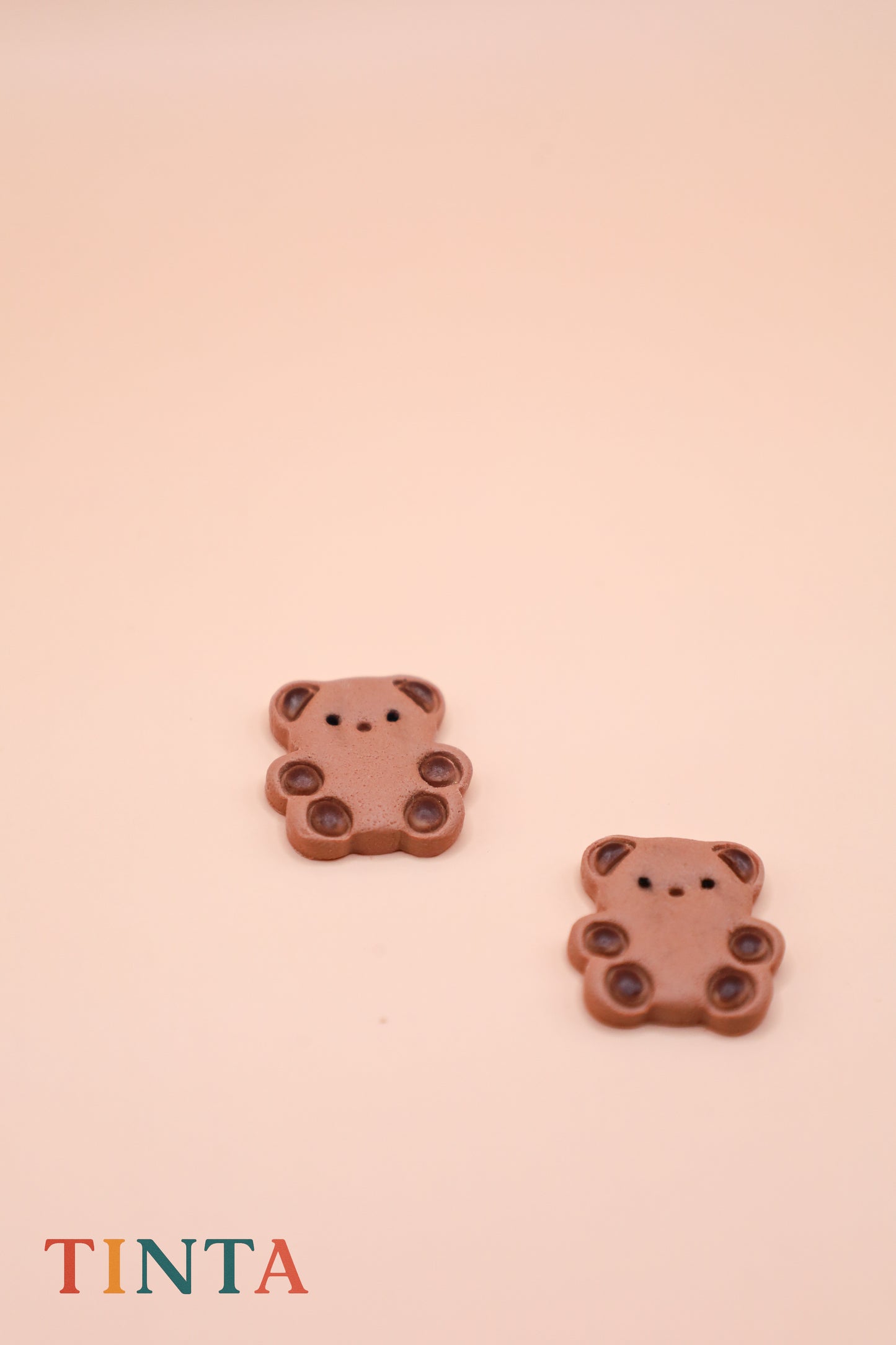Two teddy bear-shaped earrings on a pink background with 'TINTA' branding.