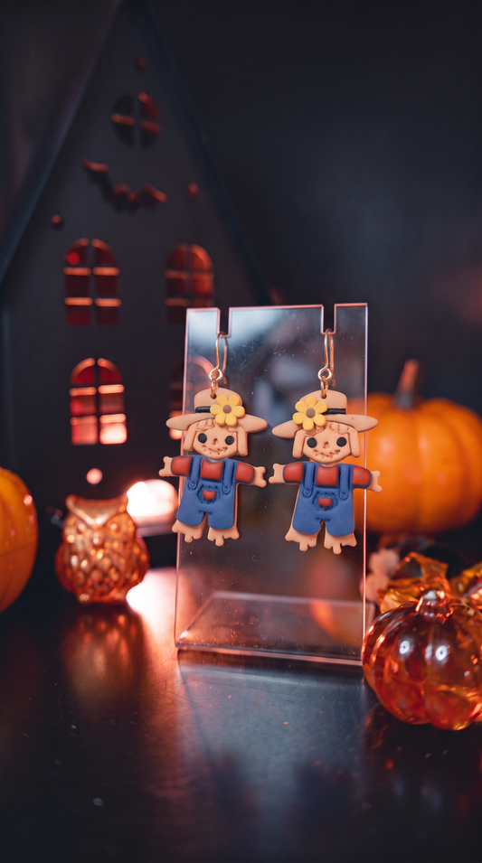 A pair of Halloween-themed scarecrow dangle earrings displayed on a clear acrylic stand. The scarecrows have tan skin, blue overalls, and straw hats with yellow flowers, set against a dark, atmospheric background with pumpkins and a house silhouette.