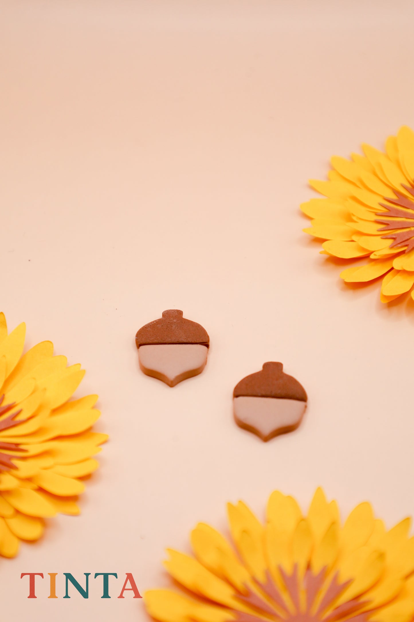 Acorn-shaped earrings with yellow flowers on a beige background, featuring the brand 'TINTA'.