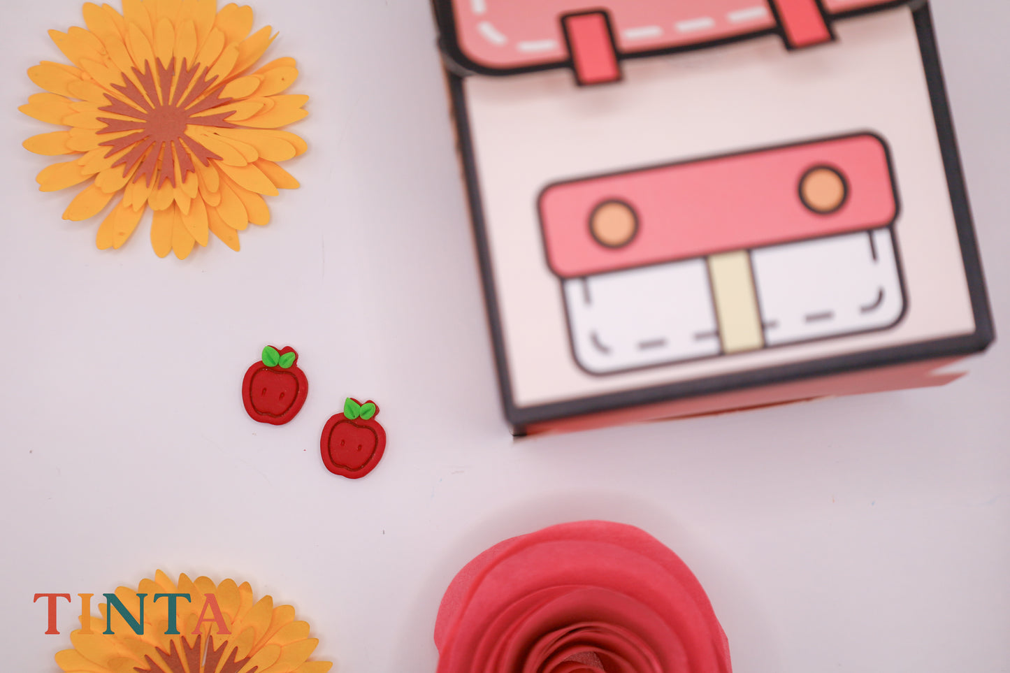 Colorful paper craft items including a pink box, red rose, and yellow flowers on a white background with 'TINTA' branding.