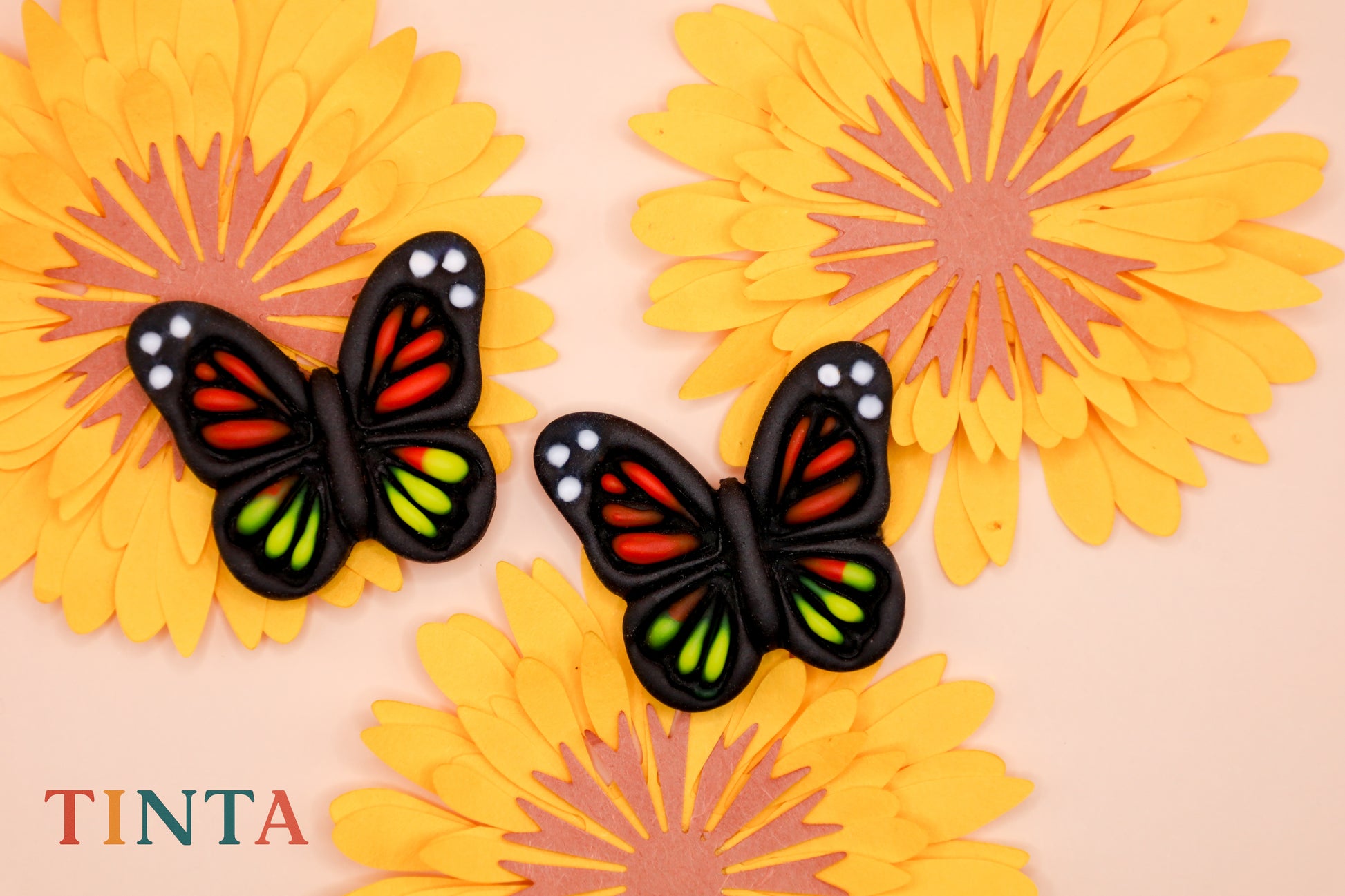 Two black butterflies with colorful wings on yellow flowers, featuring the brand 'TINTA'.