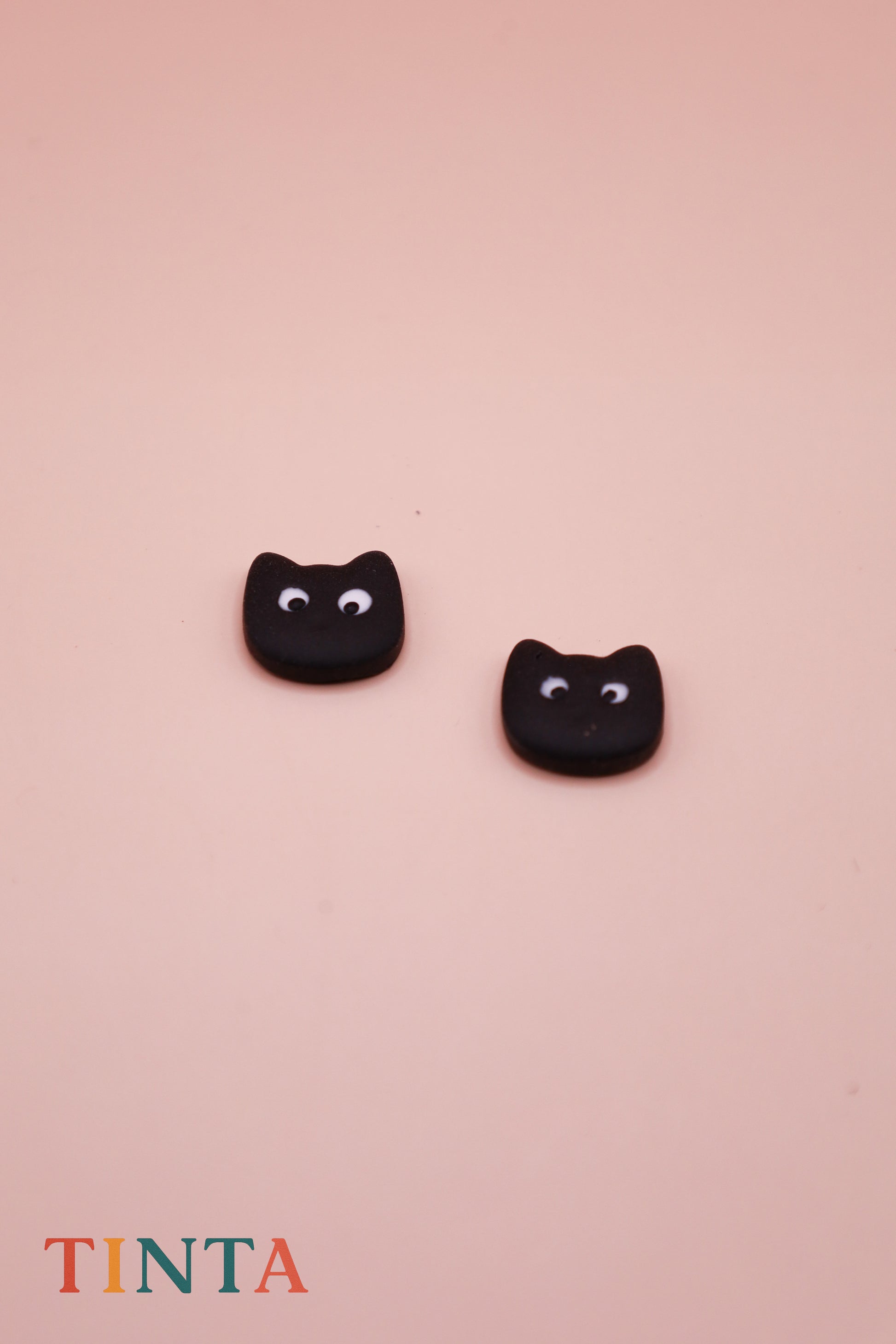 Black cat-shaped earrings on a pink background with 'TINTA' branding.