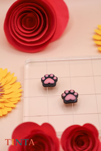 Paw print earrings on a grid with red and yellow flowers in the background, featuring the brand 'Tinta'.