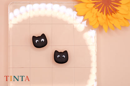 Black cat-shaped earrings on a transparent stand with a yellow flower in the background.