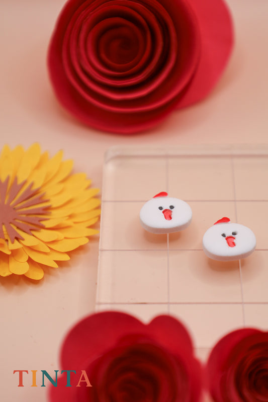 Two chicken-shaped earrings on a checkered background with red and yellow flowers.