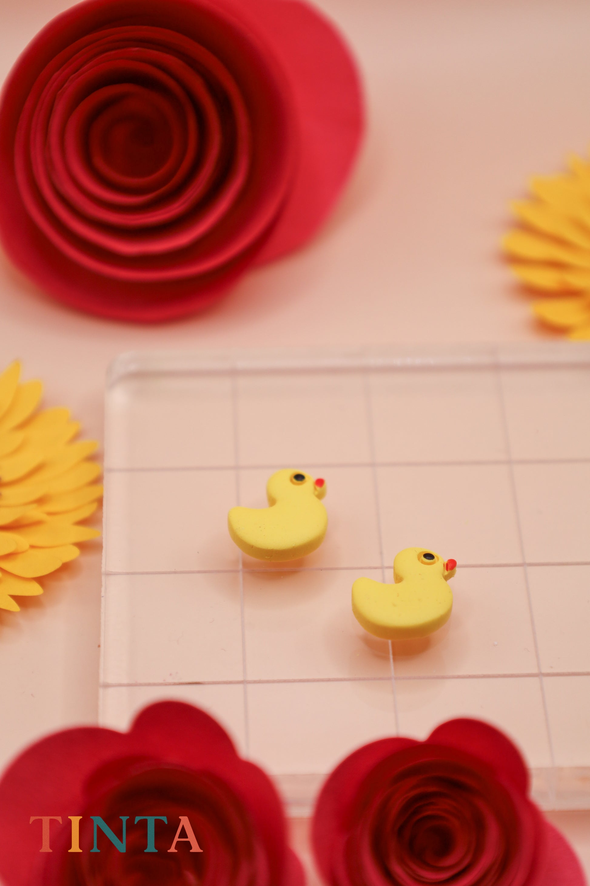 Yellow rubber duck erasers on a clear grid with red and yellow paper flowers in the background, featuring the brand 'TINTA'.