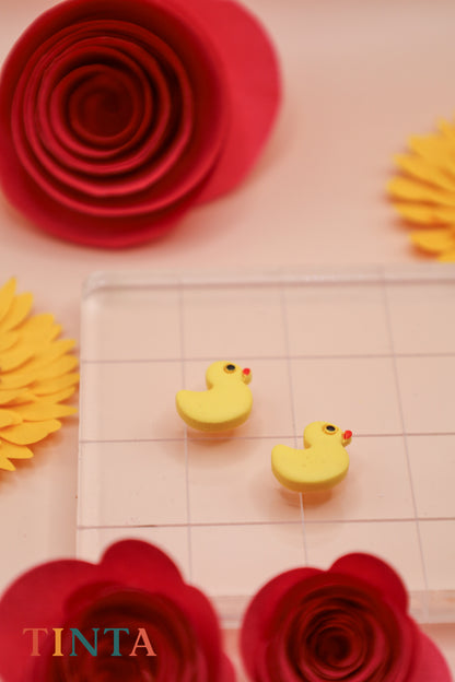 Yellow rubber duck erasers on a clear grid with red and yellow paper flowers in the background, featuring the brand 'TINTA'.
