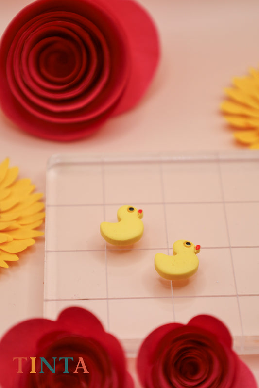 Yellow rubber duck erasers on a clear grid with red and yellow paper flowers in the background, featuring the brand 'TINTA'.