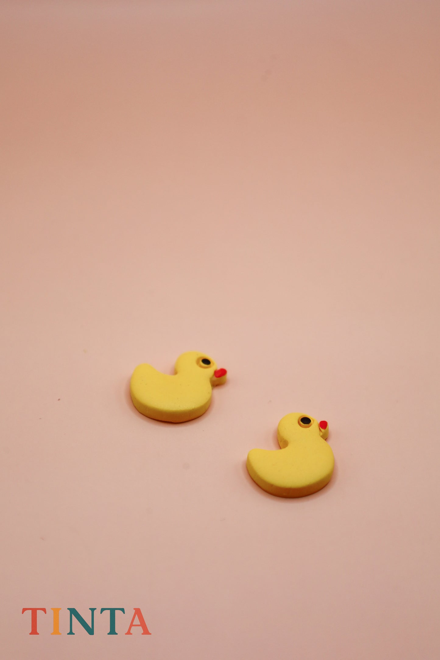 Yellow rubber duck-shaped earrings on a pink background with 'TINTA' branding.