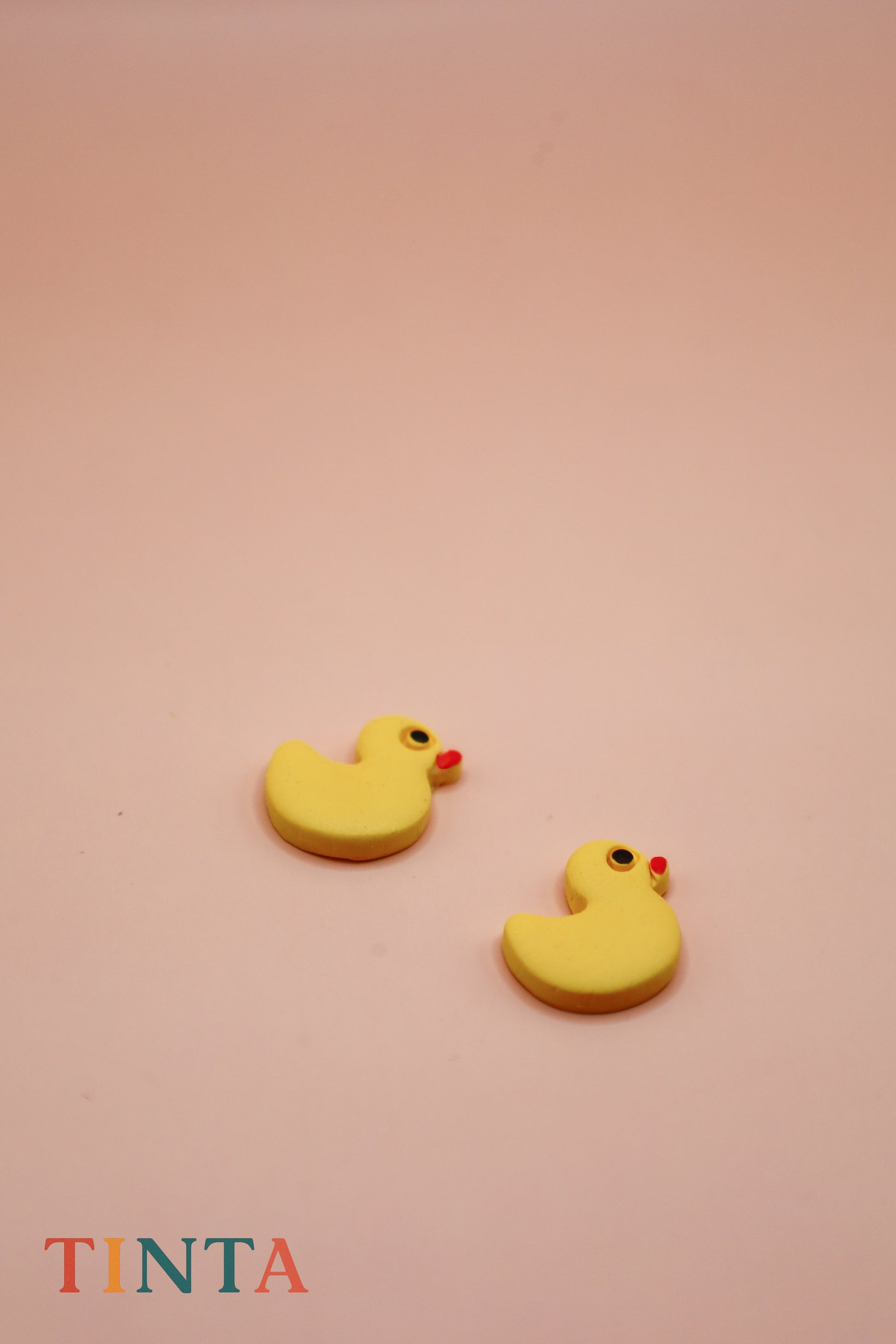 Yellow rubber duck-shaped earrings on a pink background with 'TINTA' branding.