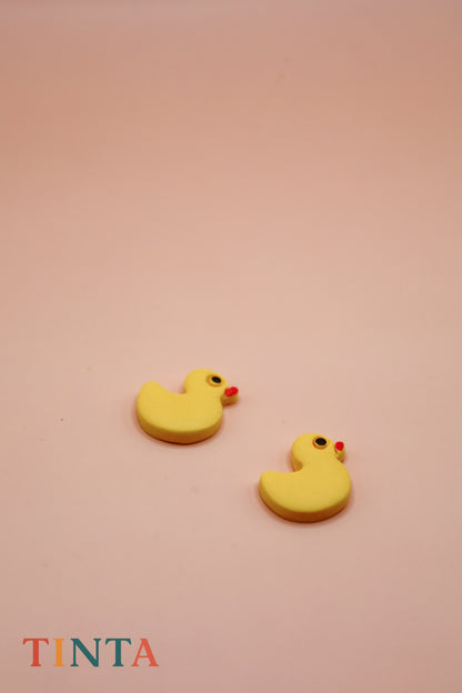 Yellow rubber duck-shaped earrings on a pink background with 'TINTA' branding.