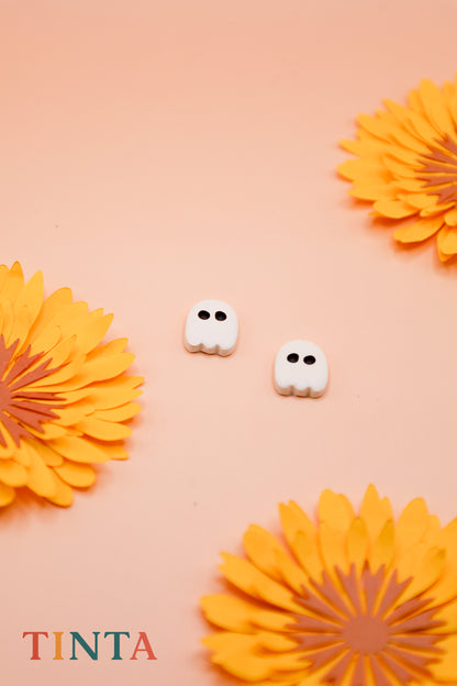 White ghost-shaped earrings on a peach background with orange flowers, featuring the brand 'TINTA'.