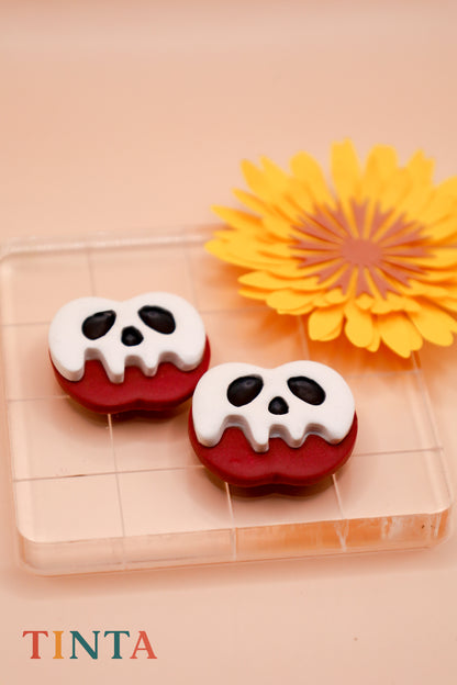 Two skull-shaped earring on a clear stand with a yellow flower in the background, featuring the brand 'TINTA'.