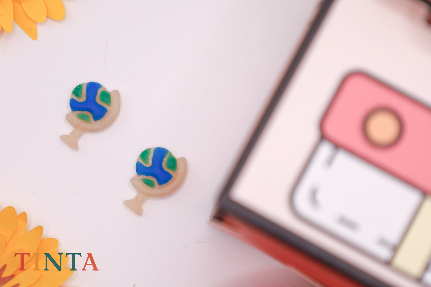 Colorful cookie-shaped earrings with a blurred product box in the background, featuring the brand 'TANTA'.