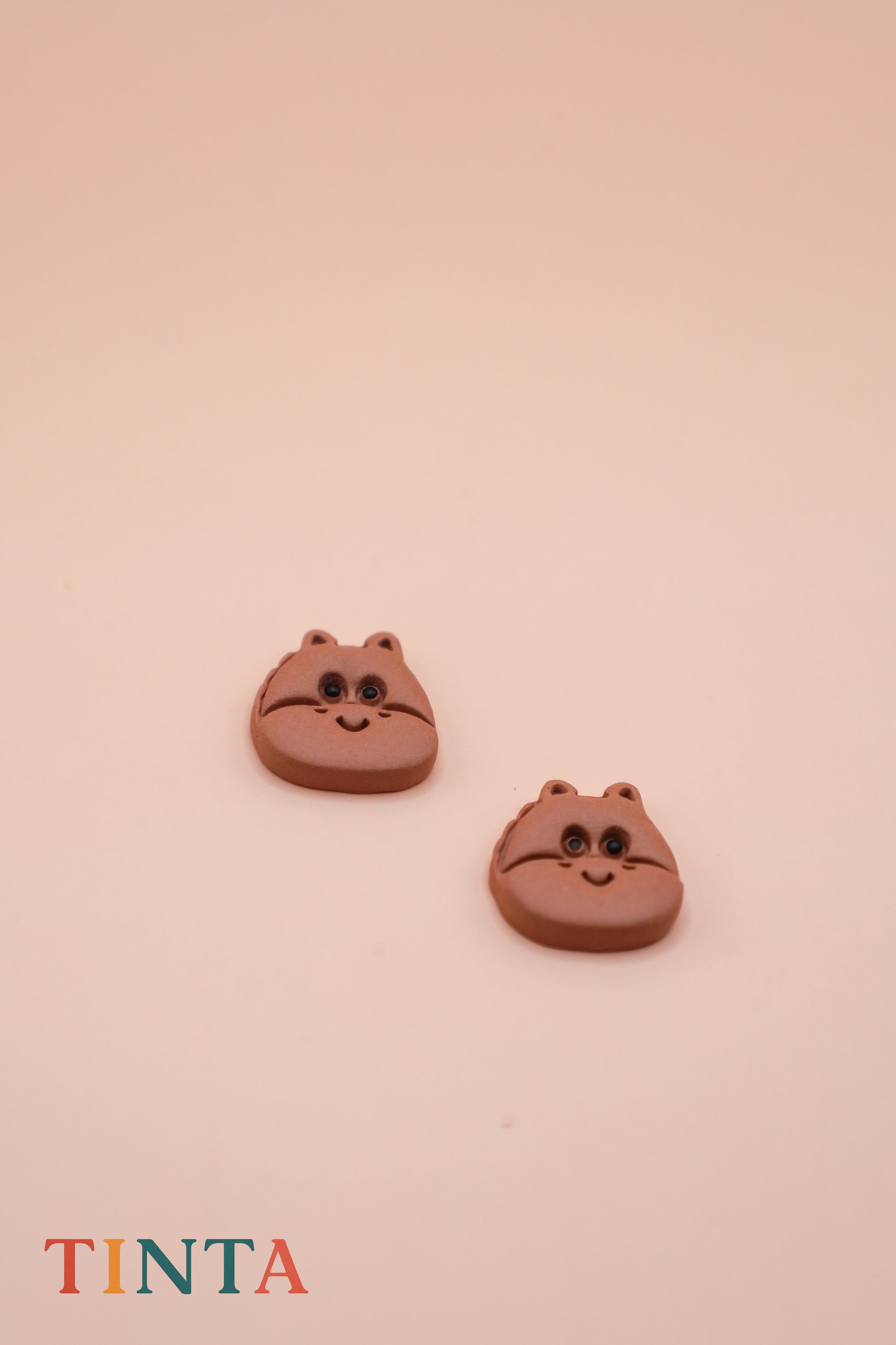 Two brown cat-shaped earrings on a beige background with 'TINTA' branding.
