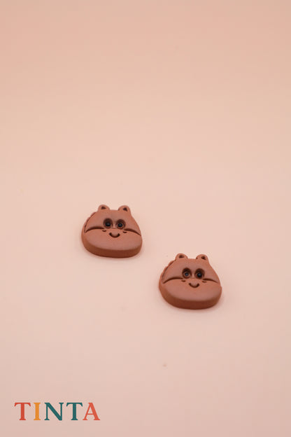 Two brown cat-shaped earrings on a beige background with 'TINTA' branding.