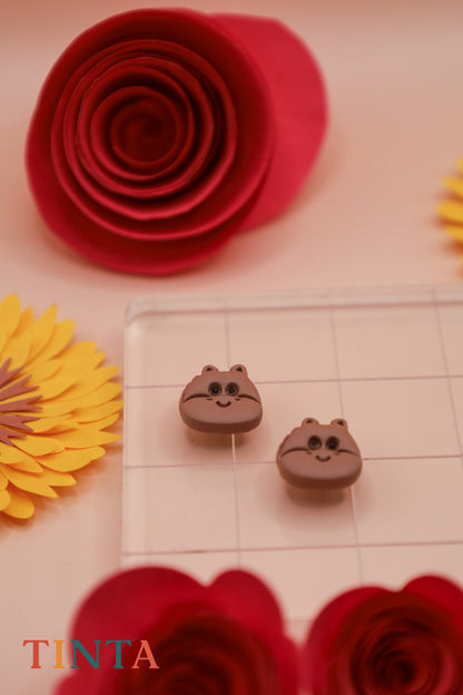 Two small brown animal-shaped earrings on a checkered surface with red and yellow flowers around.