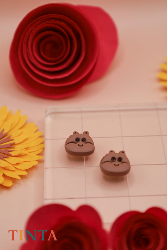 Two small brown animal-shaped earrings on a checkered surface with red and yellow flowers around.