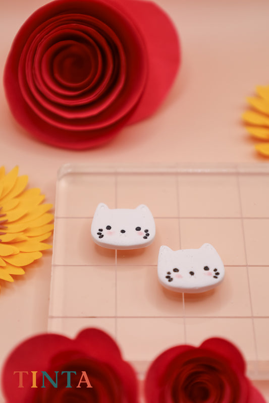 White cat-shaped objects on a checkered surface with red and yellow flowers in the background, featuring the brand 'TINTA'.