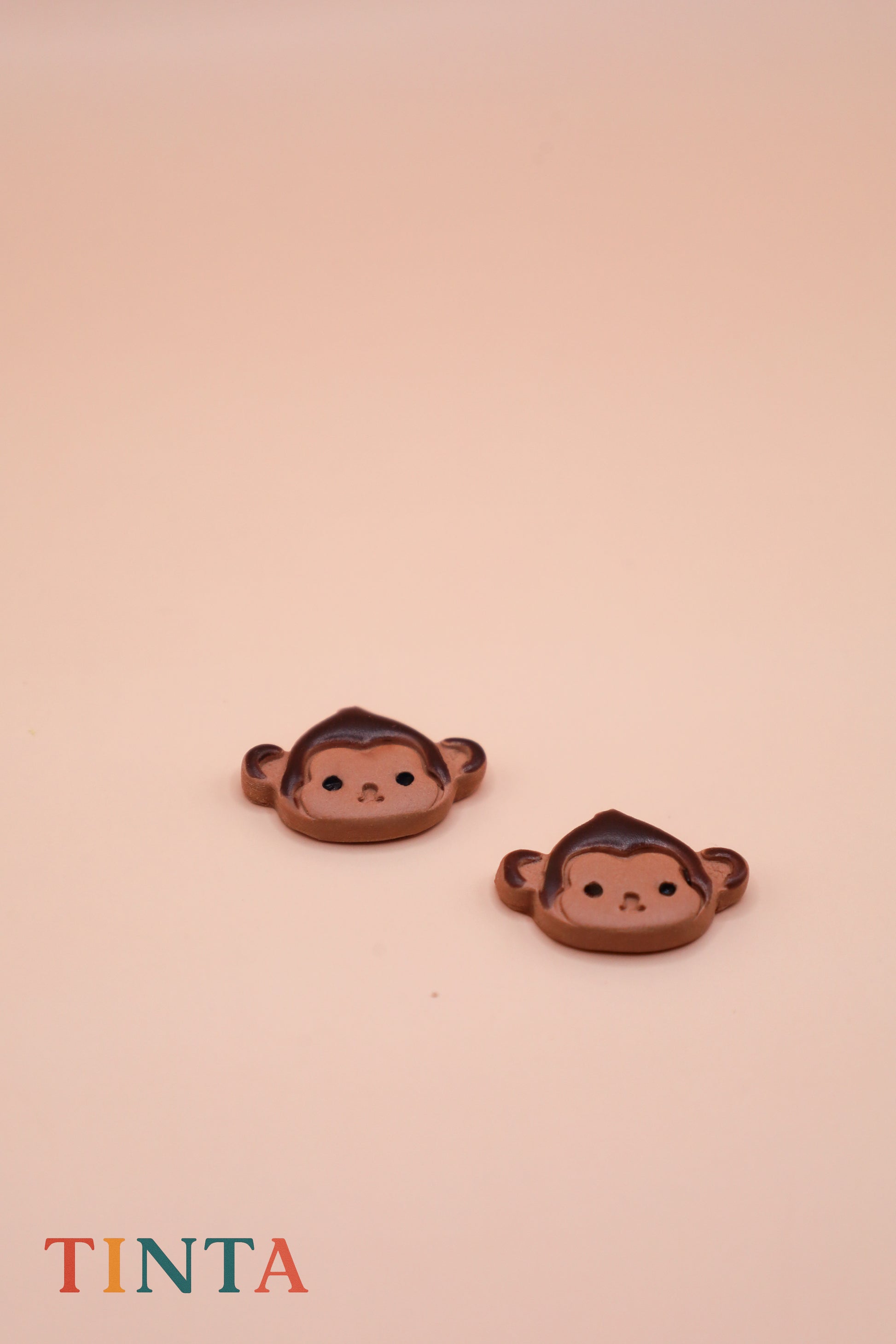 Two monkey-shaped earrings on a beige background with 'TINTA' branding.
