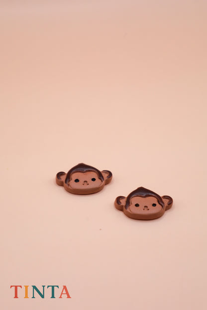 Two monkey-shaped earrings on a beige background with 'TINTA' branding.