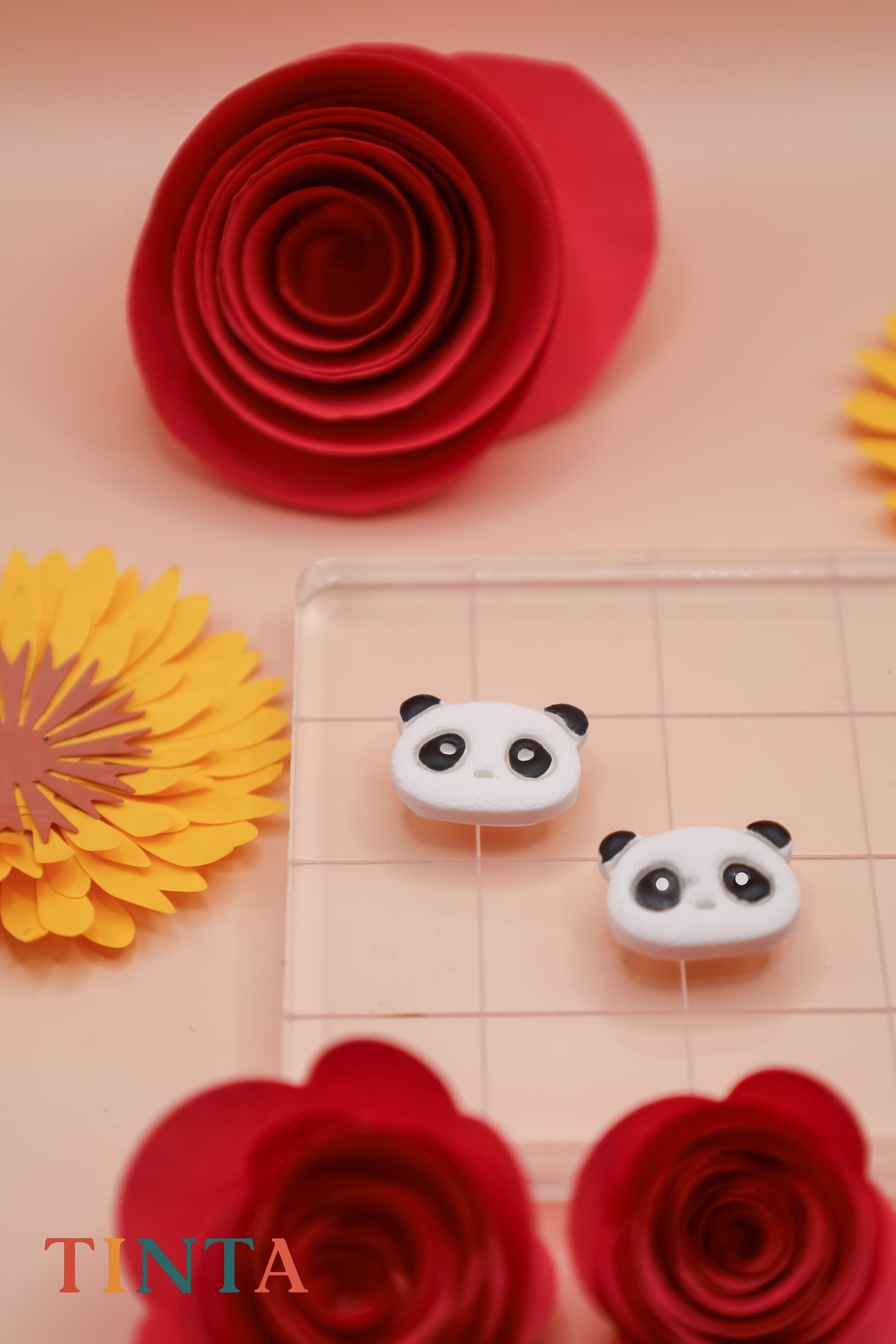 Two panda-shaped earrings on a checkered surface with red and yellow flowers in the background, featuring the brand 'TINTA'.