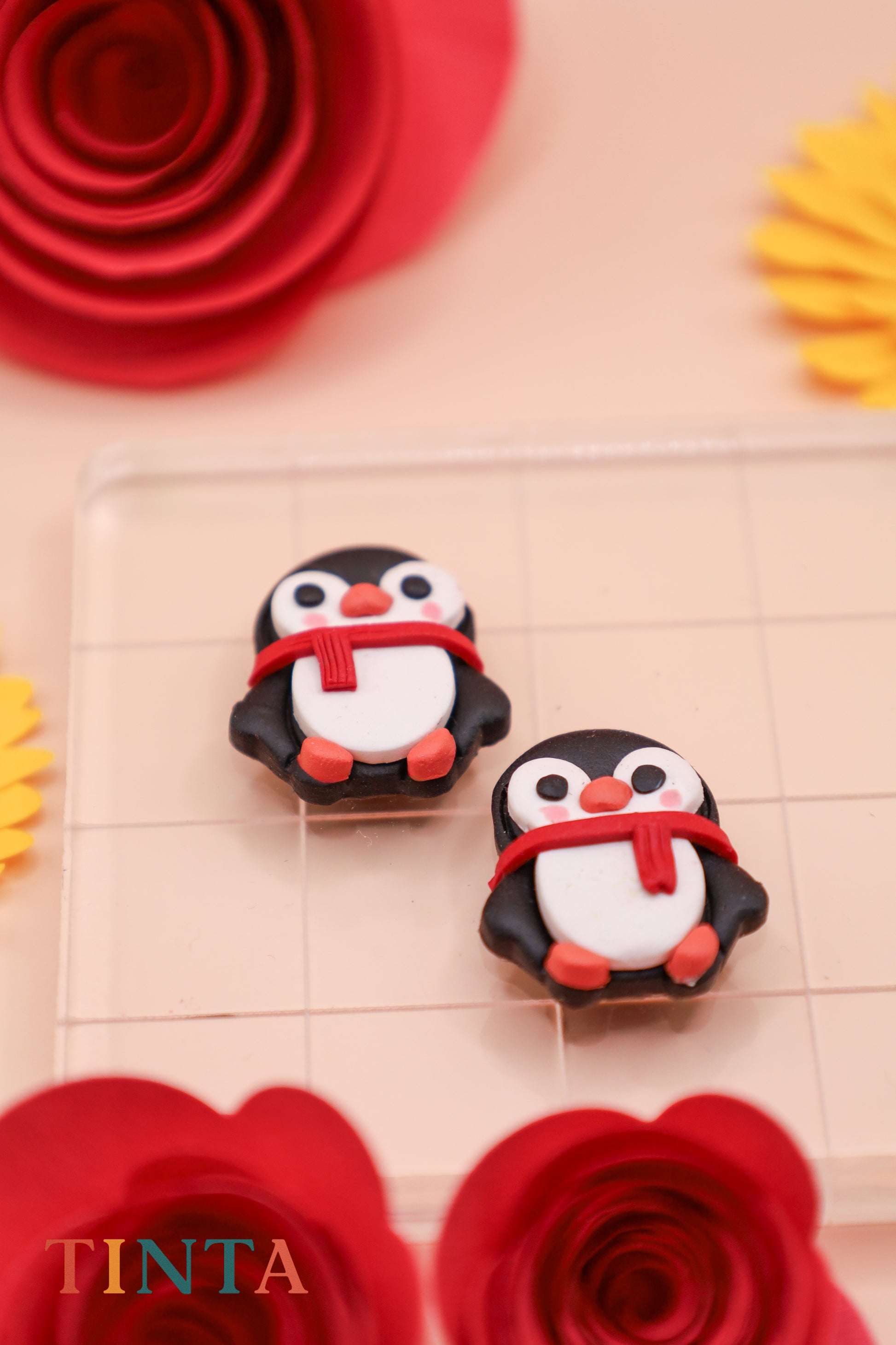 Two penguin figurines with scarves on a decorative surface with flowers and 'TINTA' branding.
