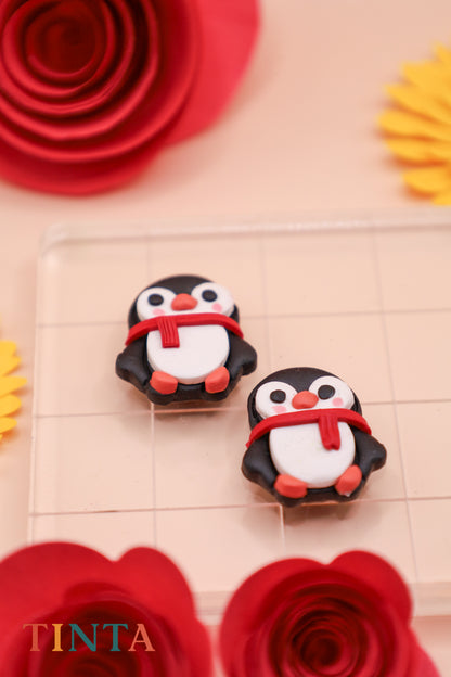 Two penguin figurines with scarves on a decorative surface with flowers and 'TINTA' branding.