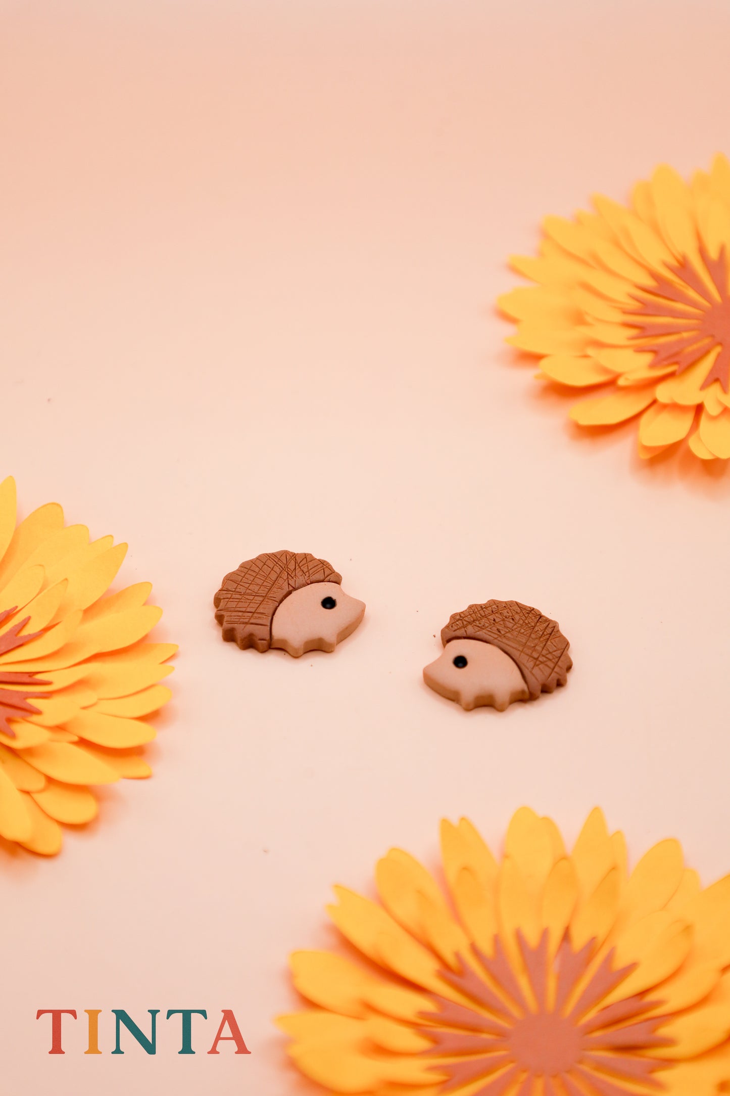 Hedgehog-shaped earrings with orange flowers on a pink background, featuring the brand 'TINTA'.