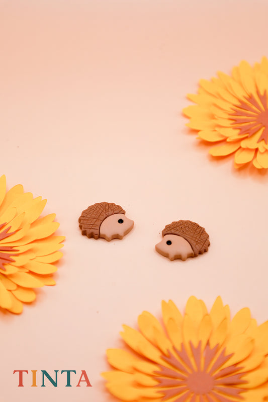 Hedgehog-shaped earrings with orange flowers on a pink background, featuring the brand 'TINTA'.
