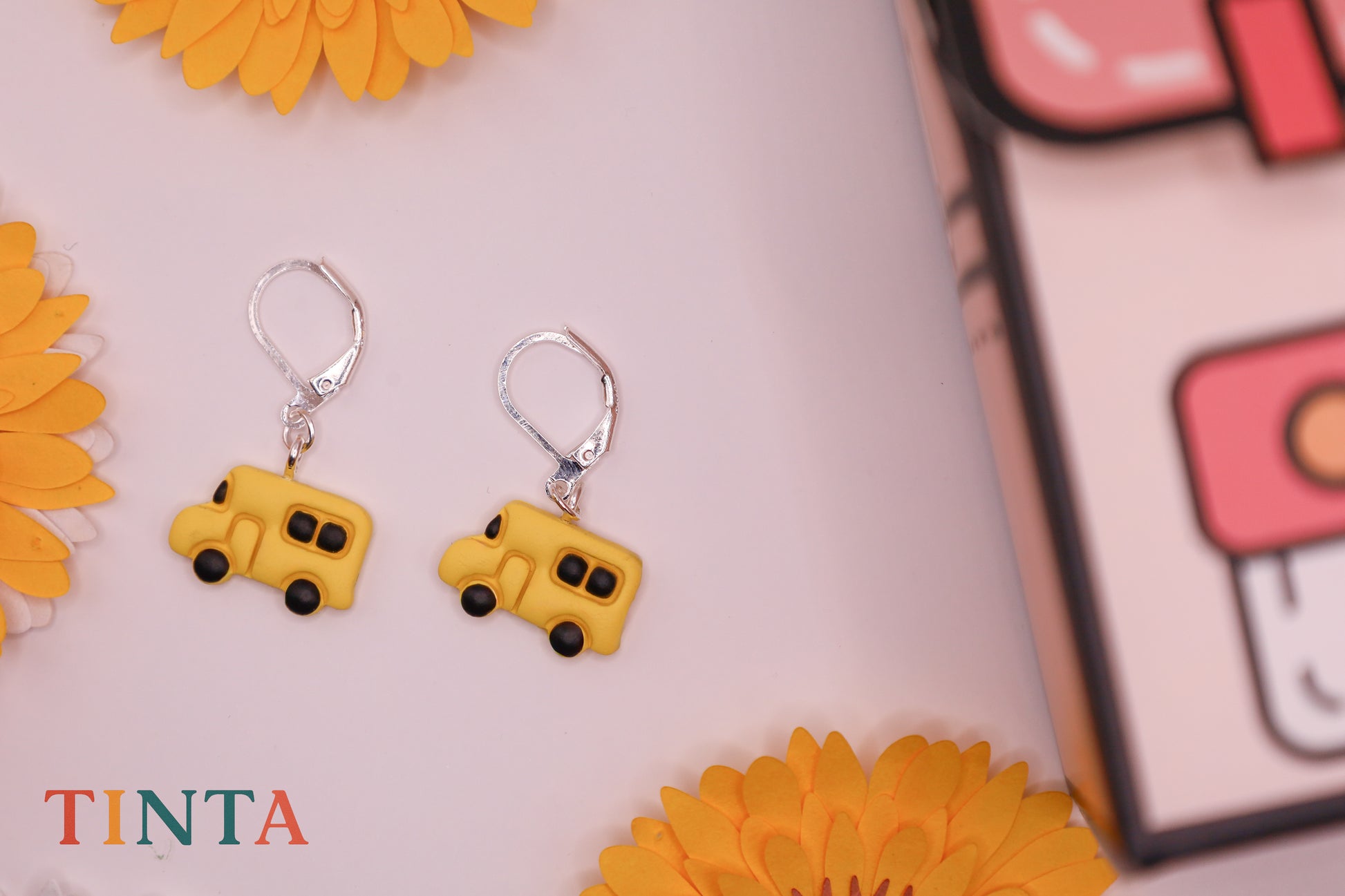 Yellow school bus-shaped earrings on a pink background with 'TINTA' branding.