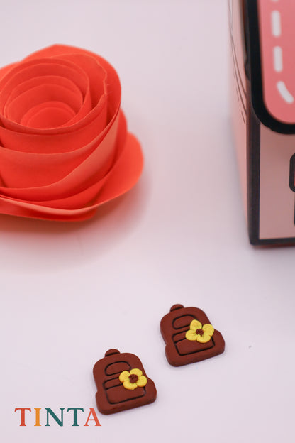 Small brown backpack-shaped earrings with yellow flowers on a white surface, next to a pink rose and a box labeled 'TINTA'.