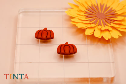 Two red pumpkin-shaped earrings on a clear stand with a yellow flower in the background, featuring the brand 'TINTA'.