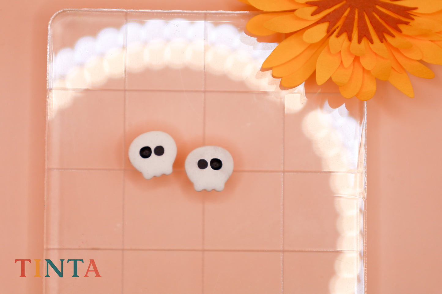 White skull-shaped earrings on a pink background with an orange flower.