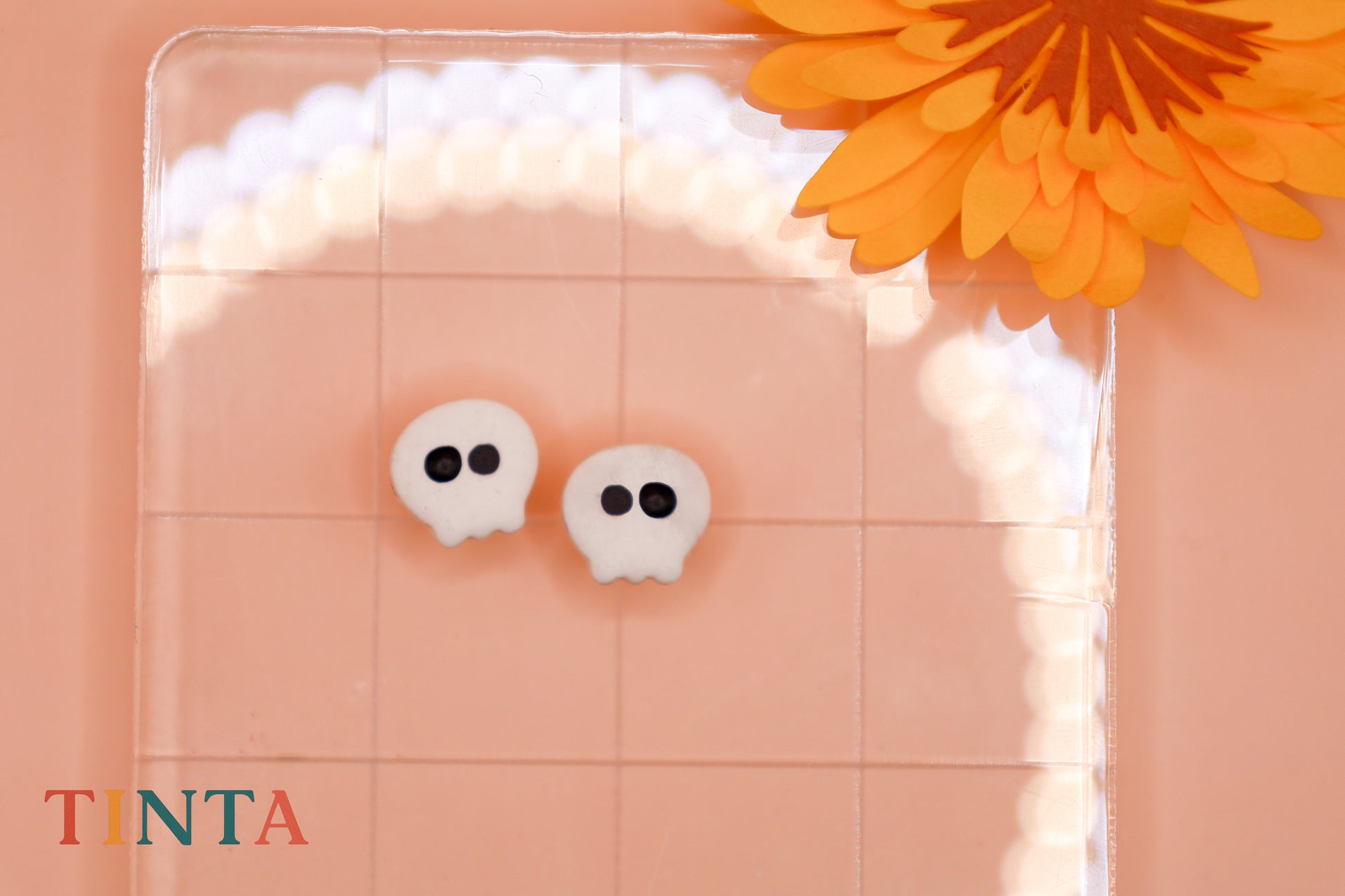 White skull-shaped earrings on a pink background with an orange flower.
