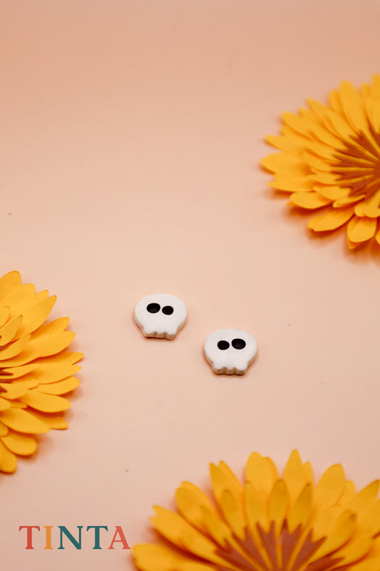 White skull-shaped earrings with black details on a beige background with orange flowers, featuring the brand 'TINTA'.