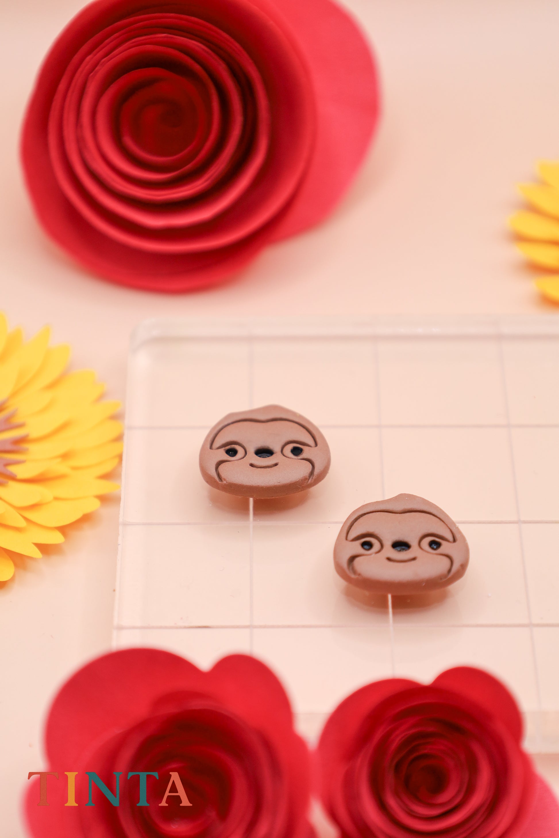 Two sloth-shaped earrings on a grid with red and yellow flowers around.