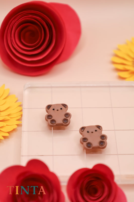 Two bear-shaped chocolate candies on a grid with red and yellow flower decorations and 'TINTA' branding.