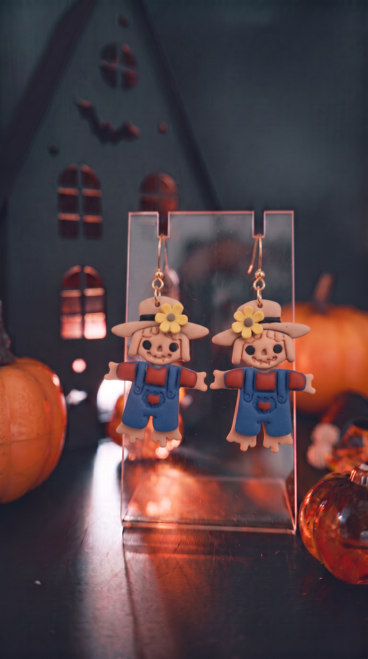 A pair of Halloween-themed scarecrow dangle earrings displayed on a clear acrylic stand. The scarecrows wear blue overalls and hats with yellow flowers, set against a dark, spooky house backdrop with pumpkins.