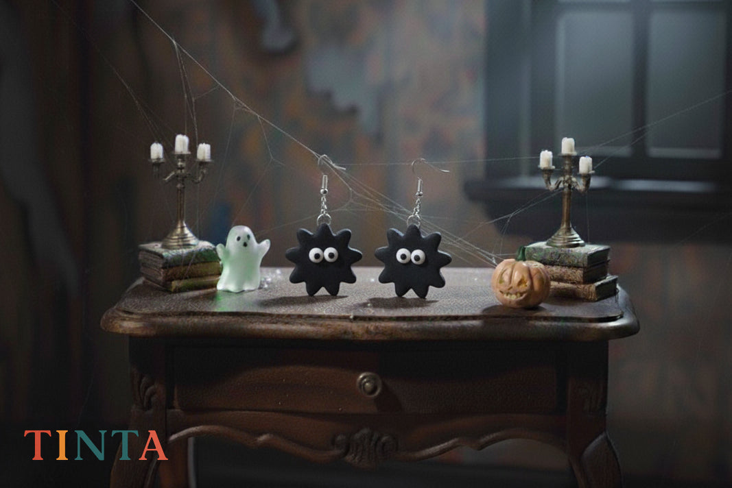 Halloween-themed earrings on a wooden table with ghost and candle decorations, featuring the brand 'Tinta'.