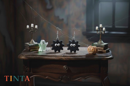Halloween-themed earrings on a wooden table with ghost and candle decorations, featuring the brand 'Tinta'.