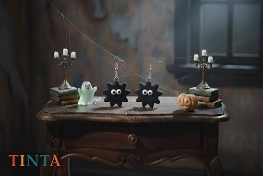 Halloween-themed earrings on a wooden table with ghost and candle decorations, featuring the brand 'Tinta'.