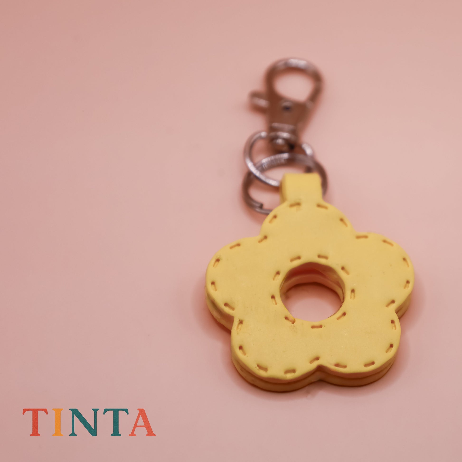 Yellow flower-shaped keychain on a pink background with 'TINTA' branding.