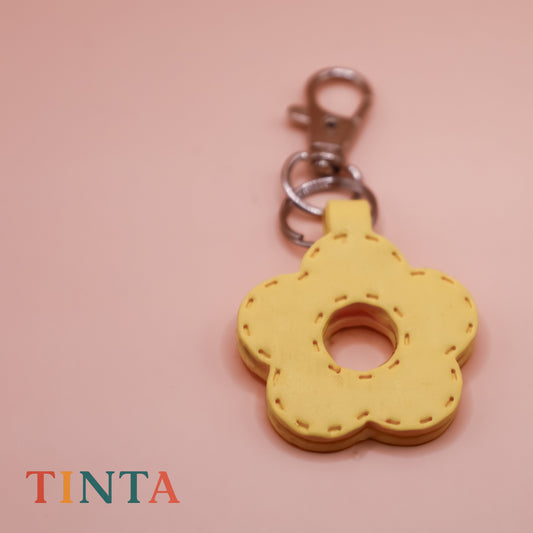 Yellow flower-shaped keychain on a pink background with 'TINTA' branding.